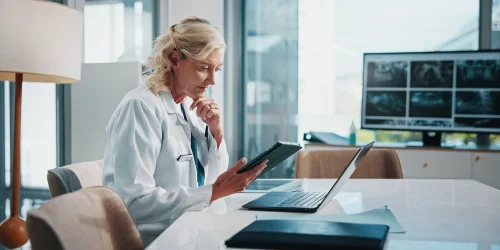 A doctor sits at a desk reviewing information on a tablet in a modern office with a monitor showing X-rays.